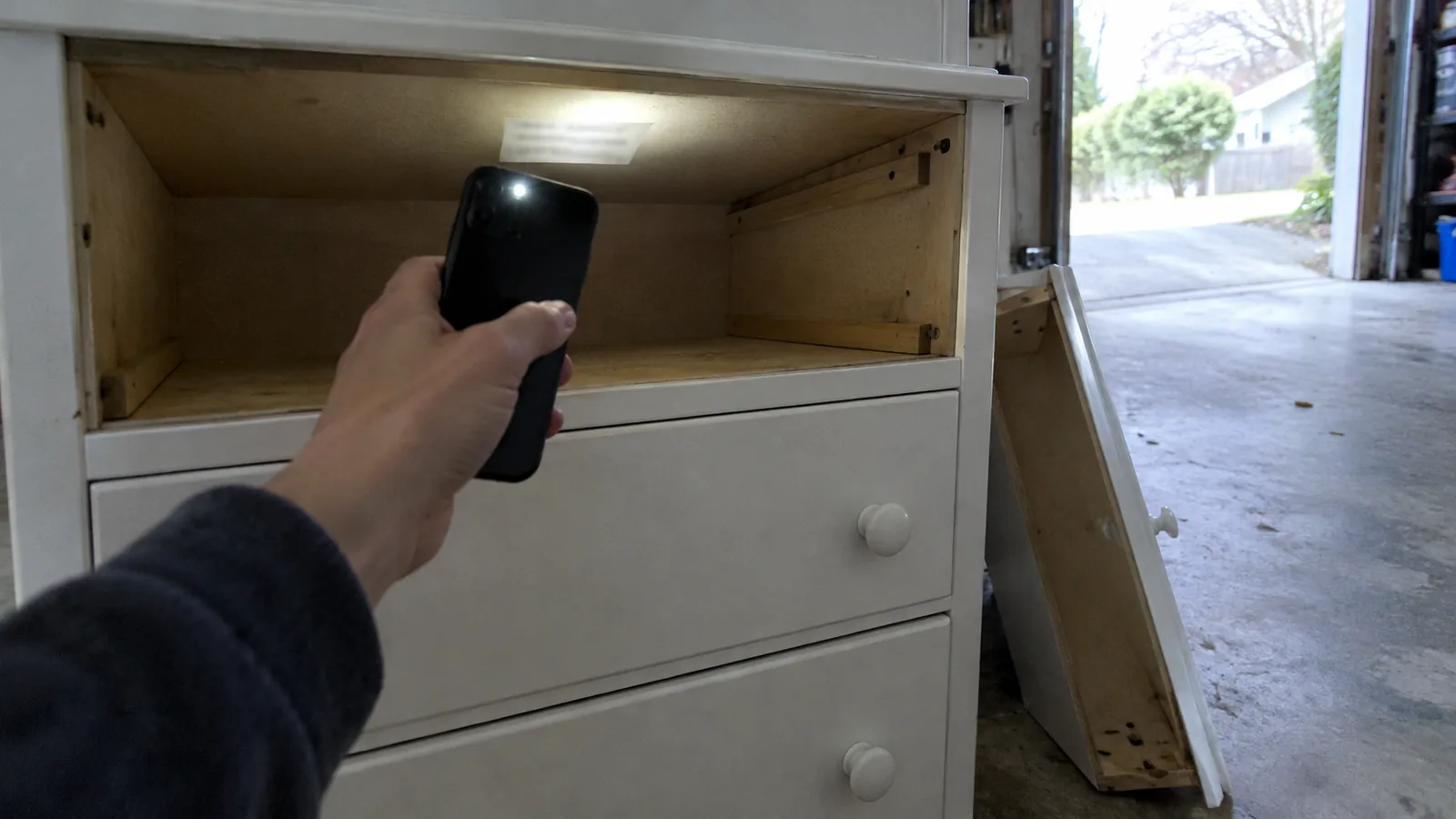 Hand shining a phone torch up into a secondhand white dresser with the top drawer removed, lighting a small label under the top panel