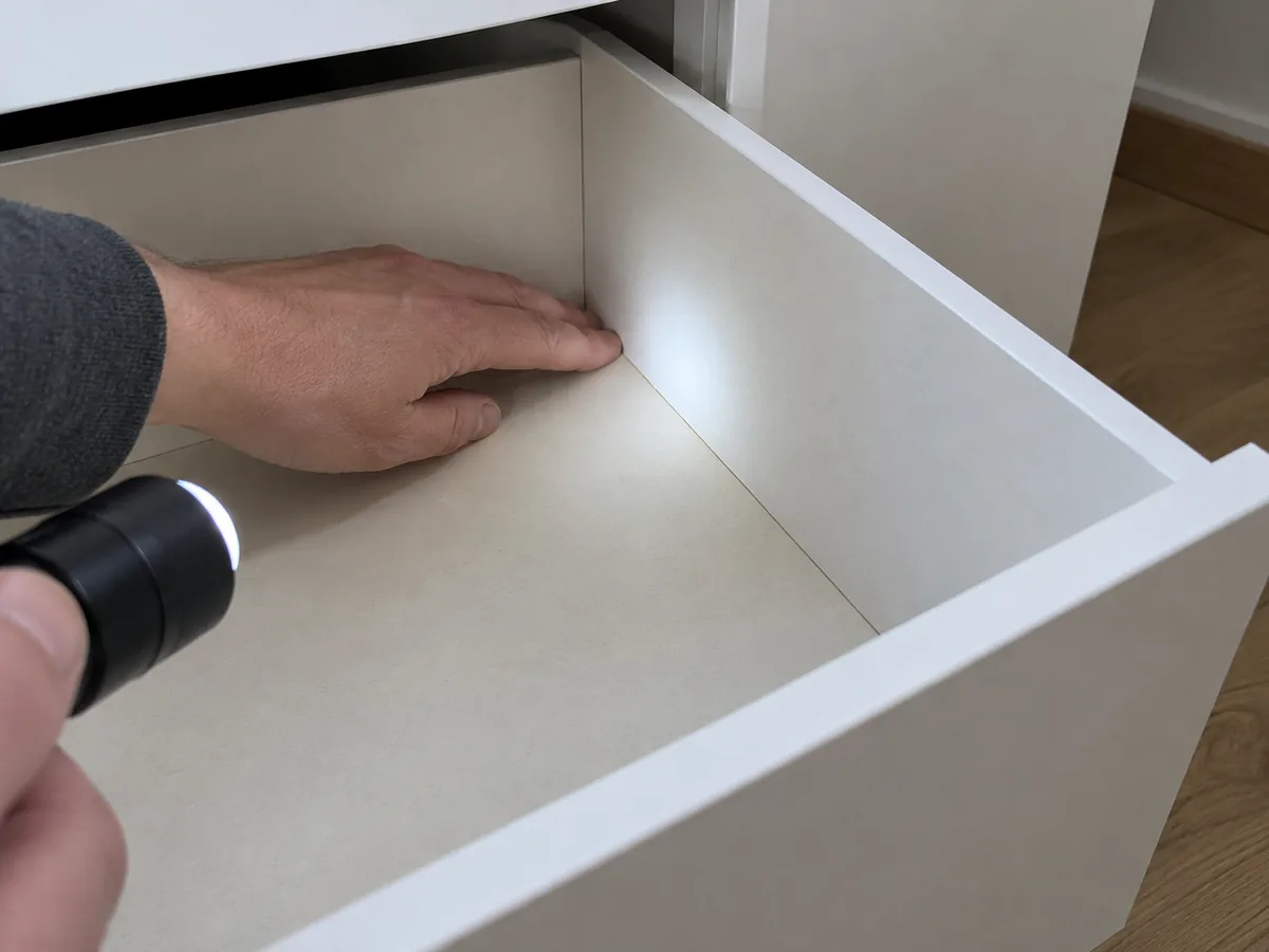 Hand and small torch inspecting the bottom-to-side joint of an empty generic white dresser drawer