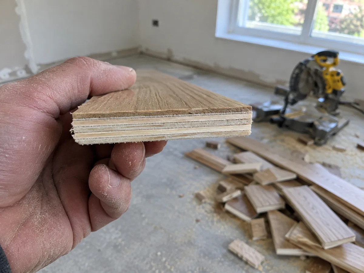 Hand holding the cut end of an engineered oak floorboard offcut showing a thin layer of oak on top of a plywood core
