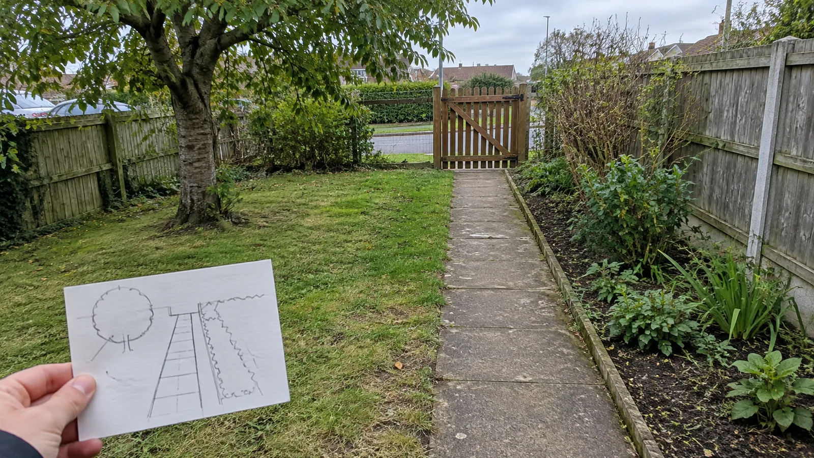 Hand holding a simple yard sketch with a path, gate, tree and planting border visible behind it