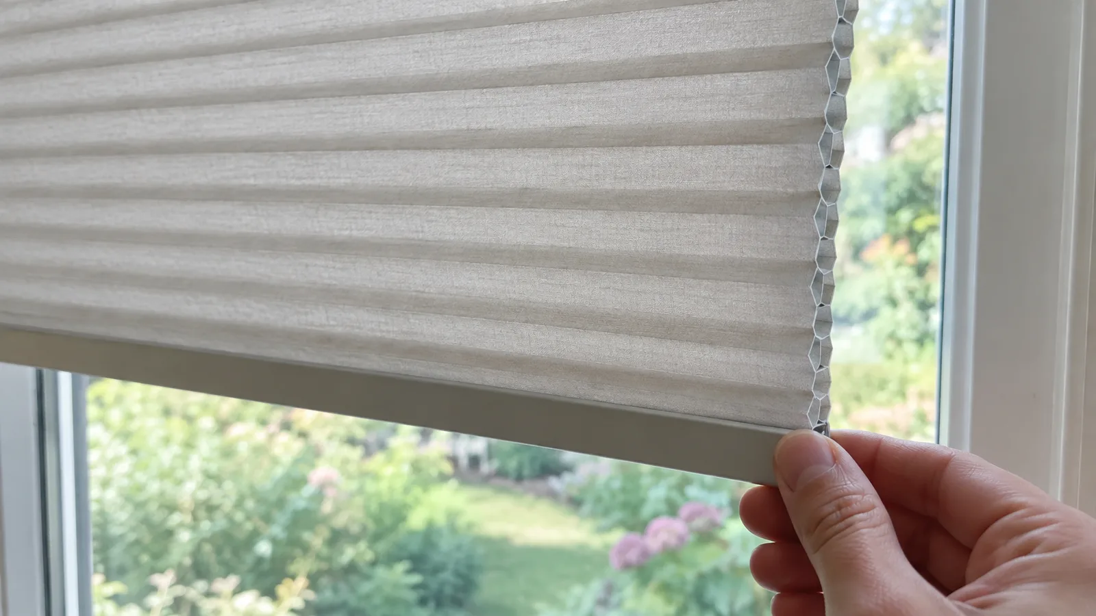 Close-up of a hand holding the corner of a cellular shade, with the full-width bottom rail below and a row of small honeycomb cells visible along the side edge