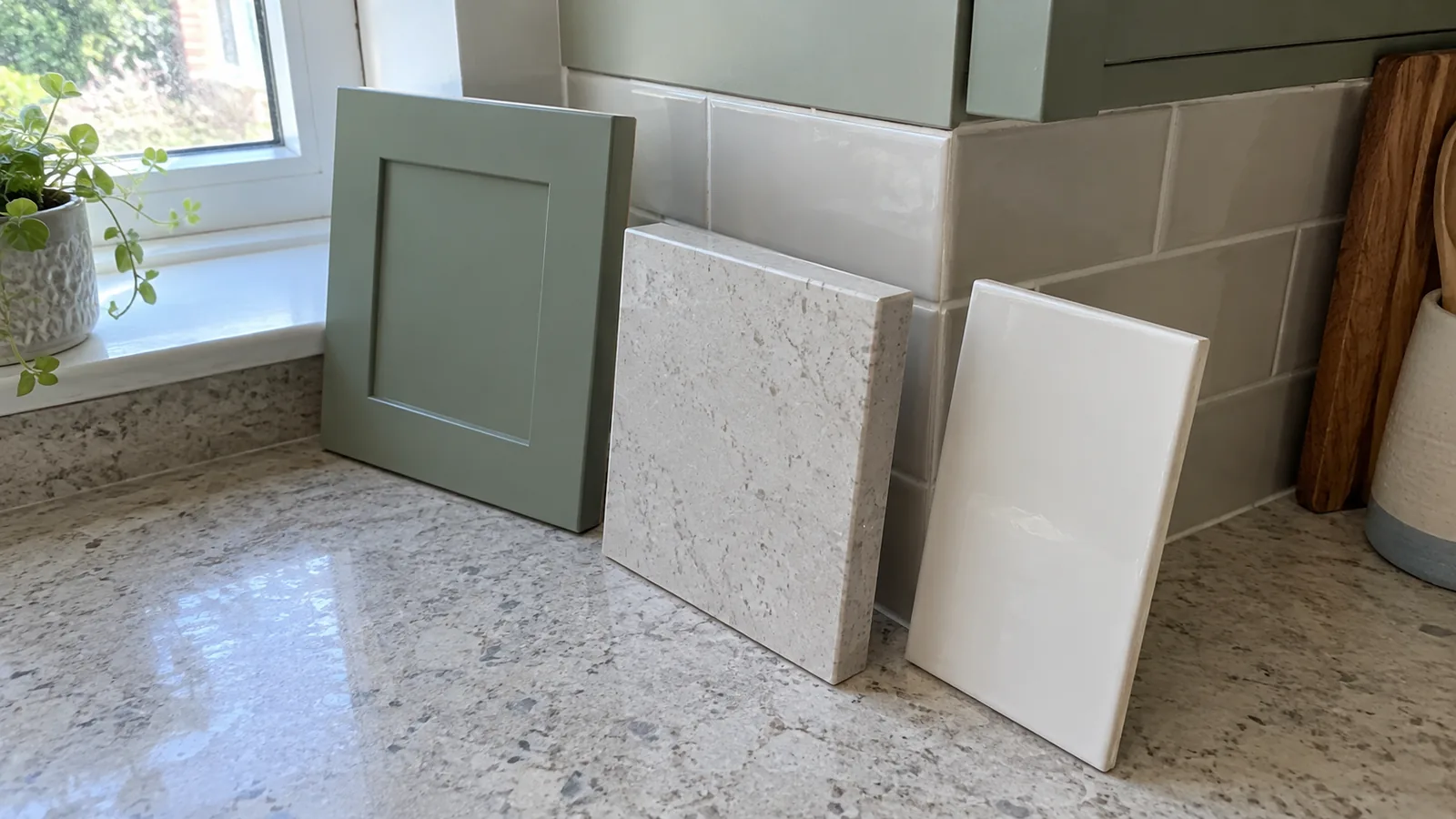 Sage cabinet-front board, pale countertop sample and white tile leaning together near a kitchen window