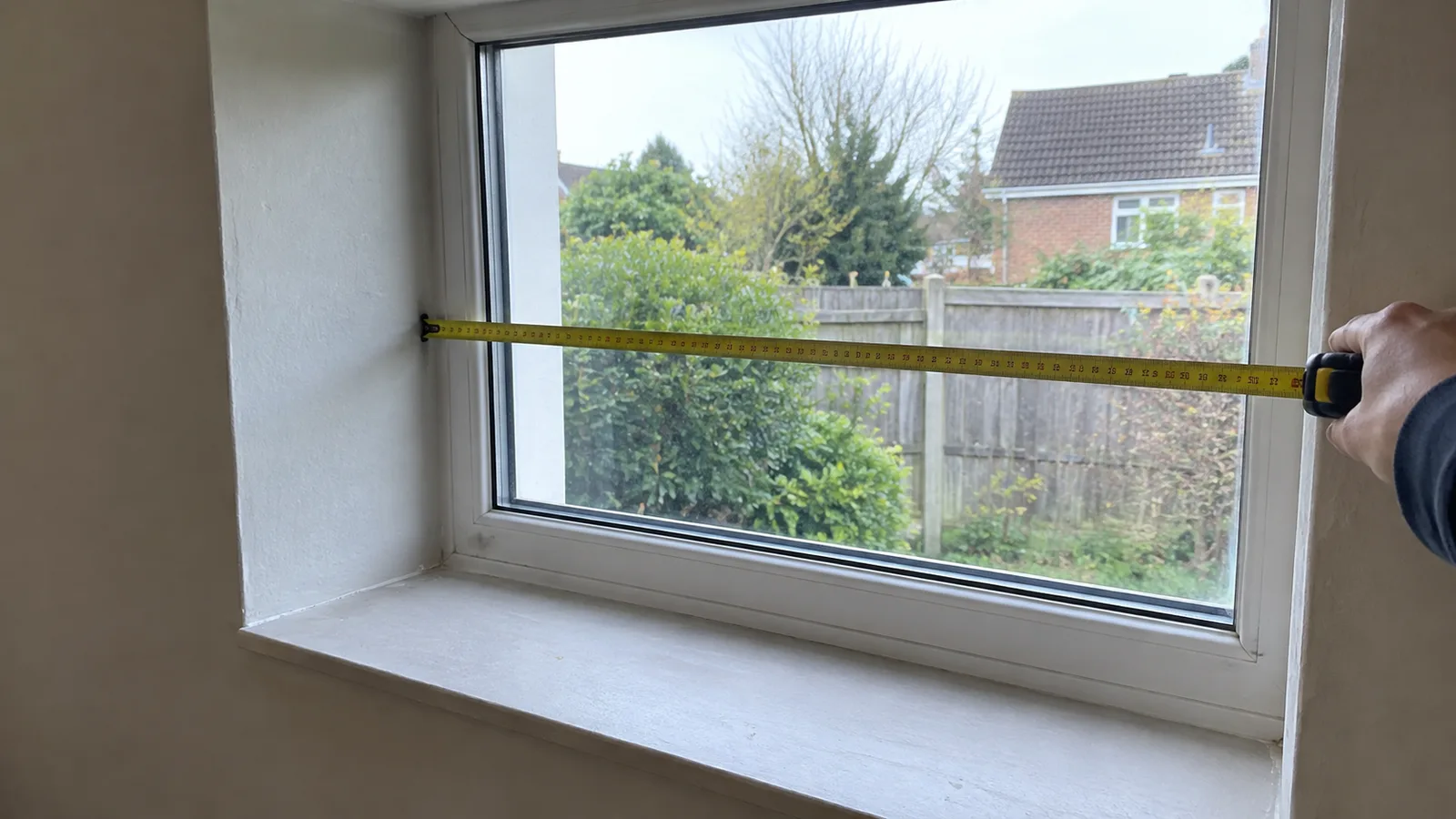 A person holding a tape measure across the width of a window frame at its top corner, with the window recess visible