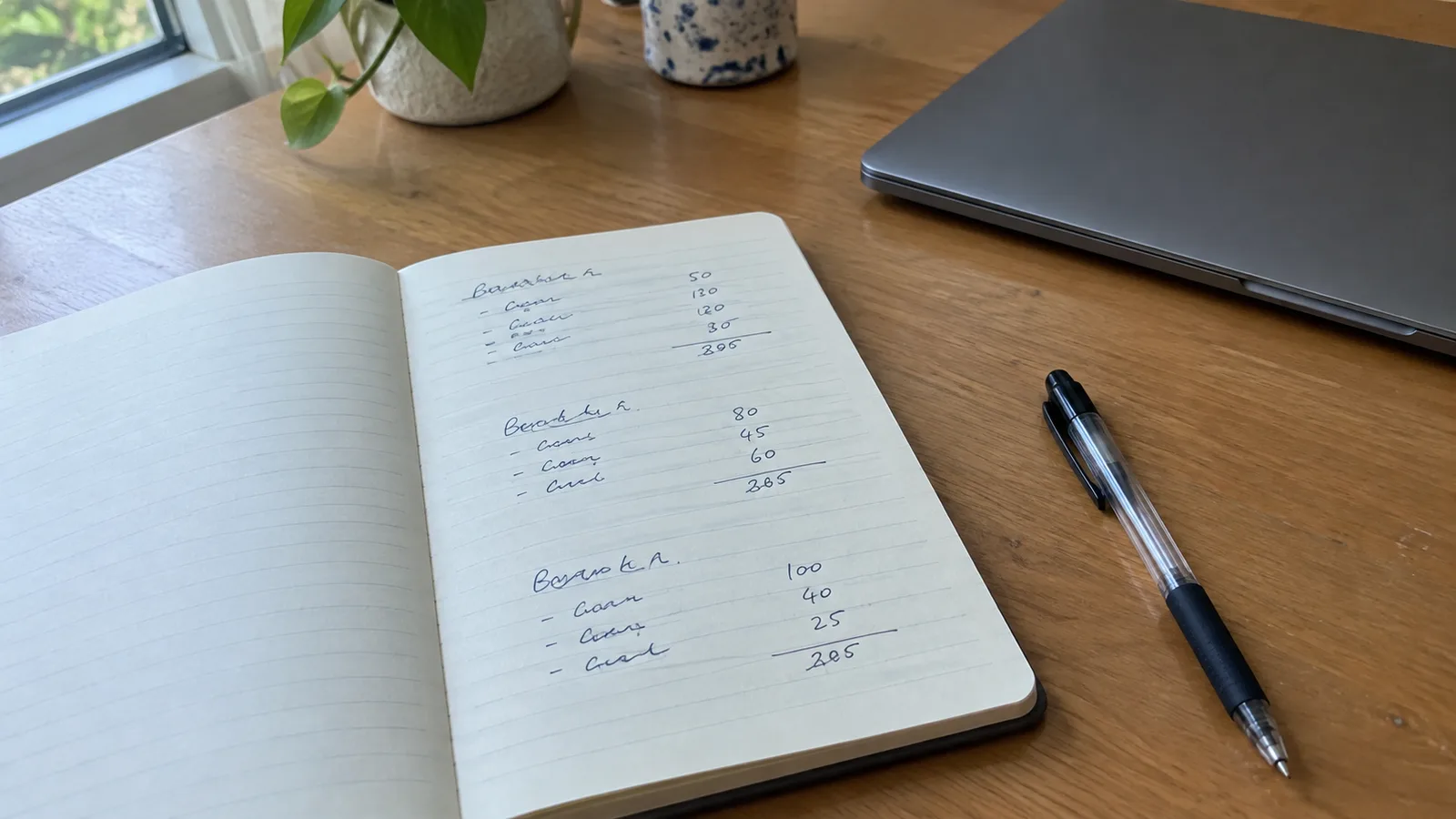 A notebook open to handwritten budget lists with totals, beside a closed laptop and a pen