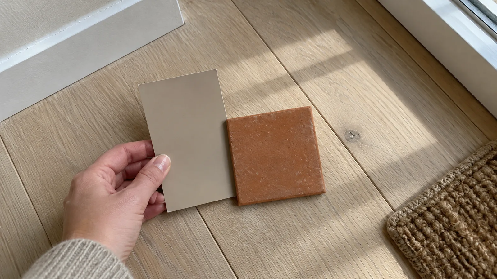 Hand holding a paint chip and a small tile sample against a pale oak floor beside a window