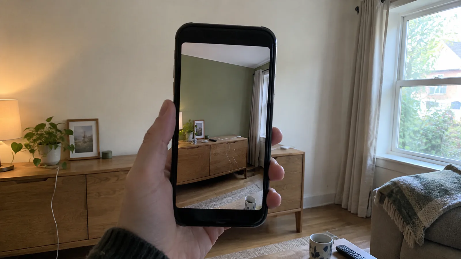 Hand holding a phone up to a living room wall, the screen showing the same wall painted a muted green through the camera