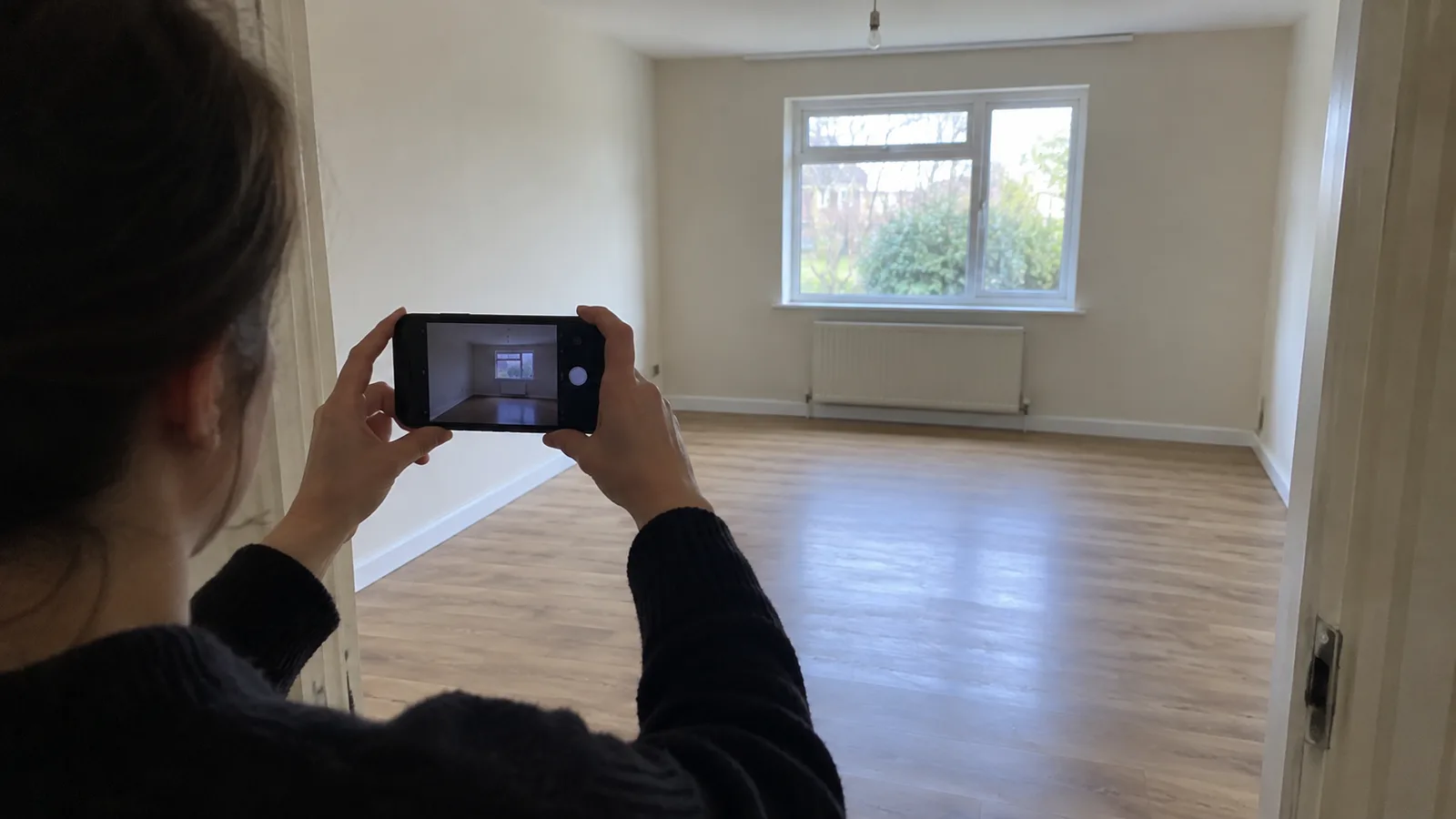 A person photographing an empty living room with a phone from the doorway before any staging is added