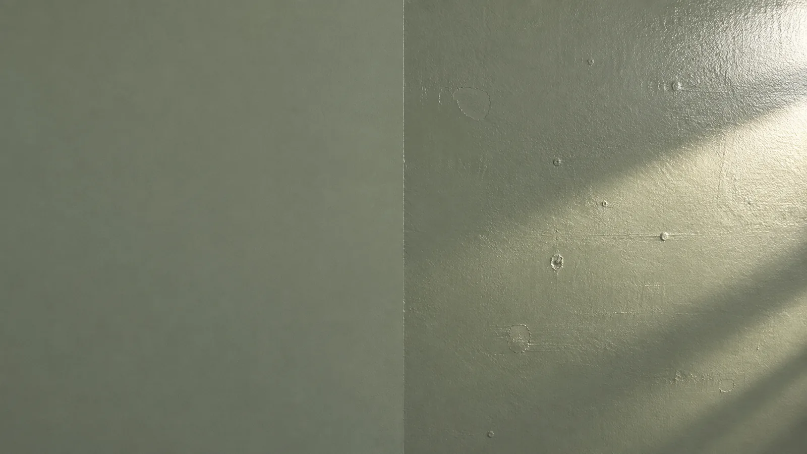 Low sunlight raking across a painted wall, showing patched dents and roller marks clearly on a satin section and barely at all on a matte section beside it