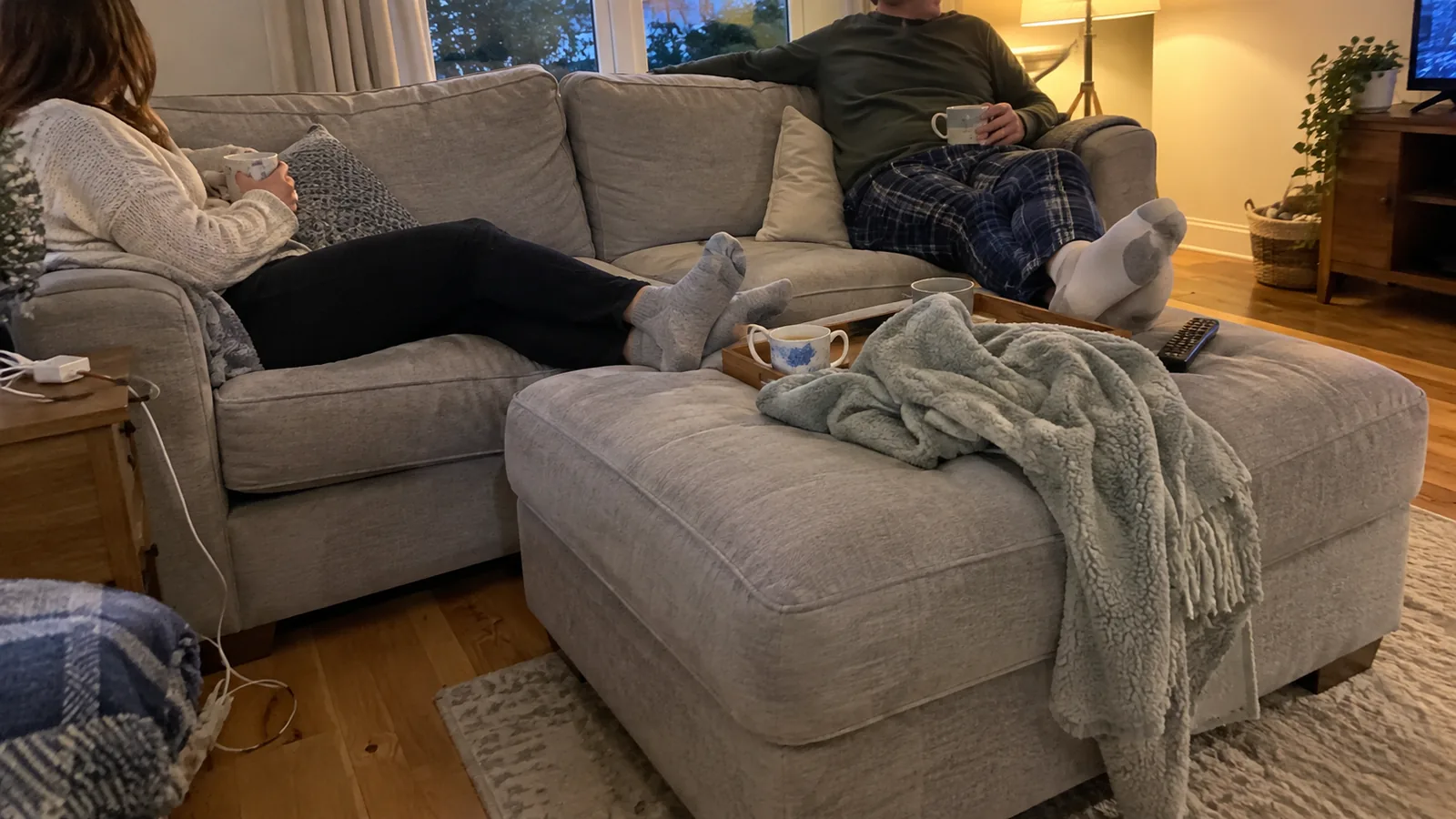 Two people at either end of a grey sofa with their feet up on one large matching ottoman, with a blanket and a mug on it