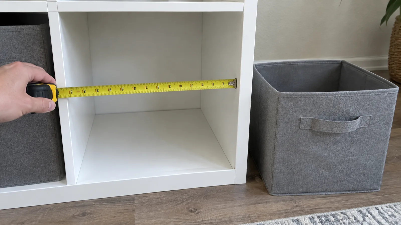 A tape measure held across one open cube of a white cube shelf, with a fabric storage bin sitting on the floor beside it to check fit
