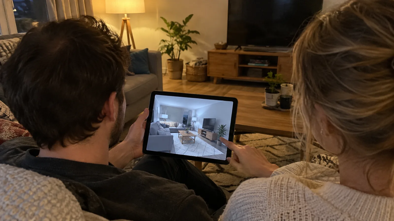 Two people sitting on a sofa looking at a 3D render of their living room on a tablet, with the real room around them